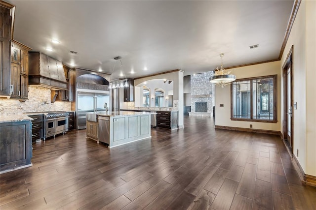 kitchen featuring decorative light fixtures, backsplash, custom exhaust hood, stainless steel appliances, and a center island with sink