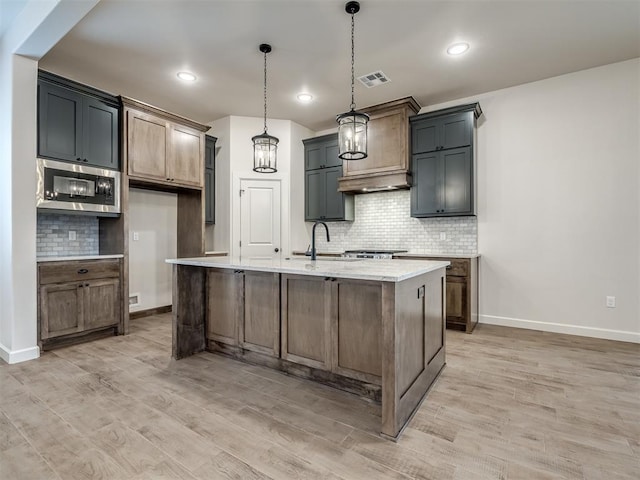 kitchen with sink, a center island with sink, light wood-type flooring, stainless steel microwave, and pendant lighting