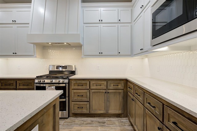 kitchen featuring white cabinetry, stainless steel appliances, tasteful backsplash, light stone countertops, and custom exhaust hood