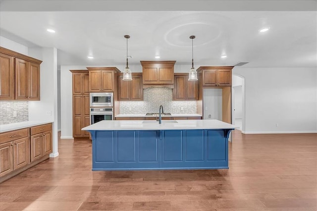 kitchen featuring hanging light fixtures, light wood-type flooring, an island with sink, stainless steel appliances, and decorative backsplash