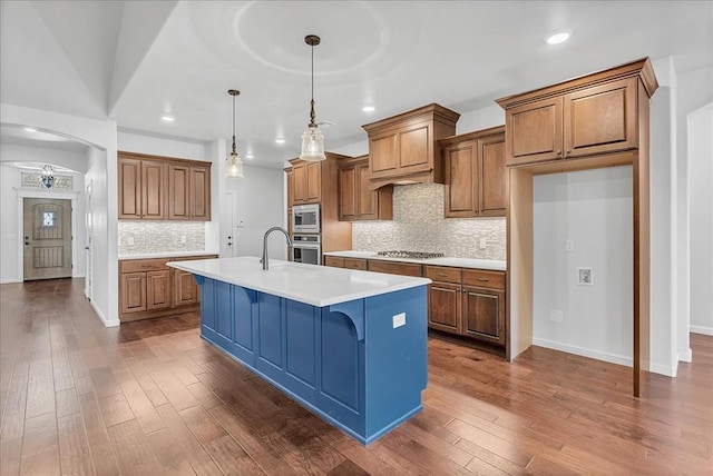 kitchen with pendant lighting, dark wood-type flooring, appliances with stainless steel finishes, a kitchen island with sink, and backsplash