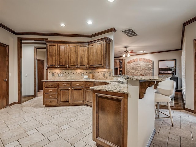 kitchen with crown molding, a breakfast bar, backsplash, light stone counters, and kitchen peninsula