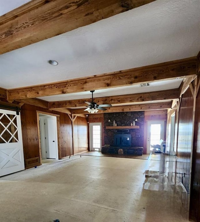 unfurnished living room with beamed ceiling, ceiling fan, a wood stove, and wooden walls