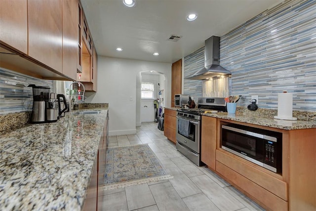 kitchen with wall chimney exhaust hood, sink, light stone counters, appliances with stainless steel finishes, and decorative backsplash