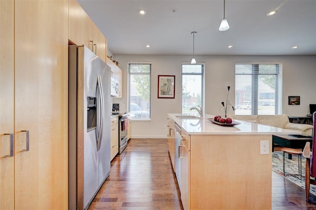 kitchen with appliances with stainless steel finishes, pendant lighting, light brown cabinetry, wood-type flooring, and a kitchen island with sink