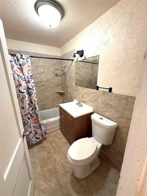 bathroom with tile walls, a textured wall, toilet, shower / bath combo with shower curtain, and a textured ceiling