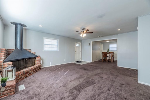 unfurnished living room with dark colored carpet, a wood stove, a wealth of natural light, and ceiling fan