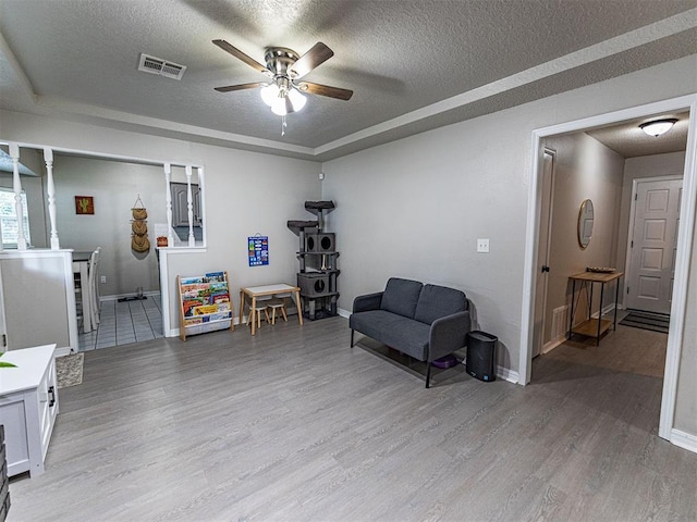 living area with ceiling fan, a raised ceiling, a textured ceiling, and light wood-type flooring