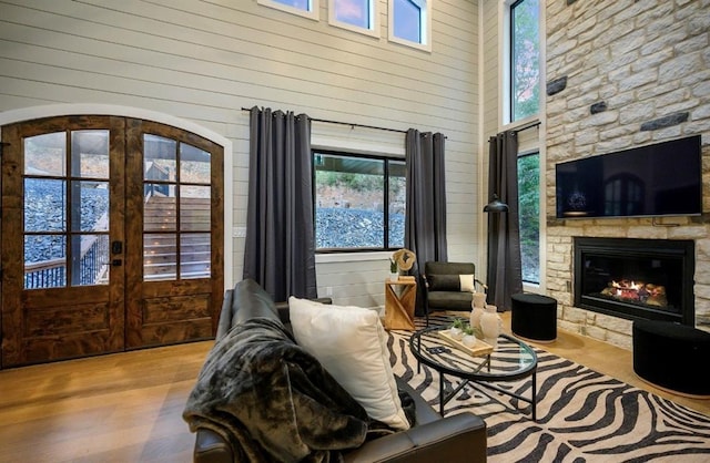 living room featuring a high ceiling, a stone fireplace, light wood-type flooring, french doors, and wood walls