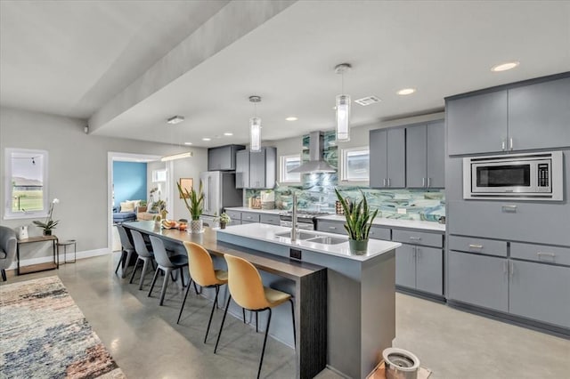 kitchen with gray cabinets, pendant lighting, stainless steel appliances, a kitchen island with sink, and wall chimney range hood