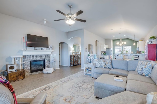 living room with lofted ceiling, ceiling fan with notable chandelier, light hardwood / wood-style floors, and a brick fireplace