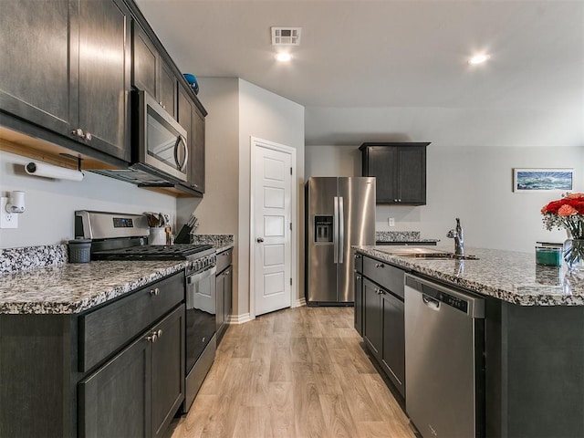 kitchen featuring sink, stainless steel appliances, light stone counters, an island with sink, and light wood-type flooring