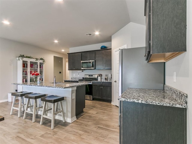 kitchen featuring a breakfast bar area, stainless steel appliances, light stone counters, light hardwood / wood-style floors, and an island with sink