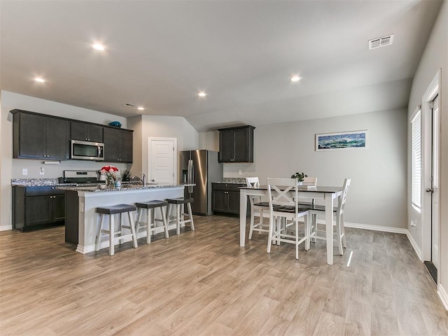 kitchen with appliances with stainless steel finishes, a breakfast bar area, dark stone counters, a center island with sink, and light wood-type flooring