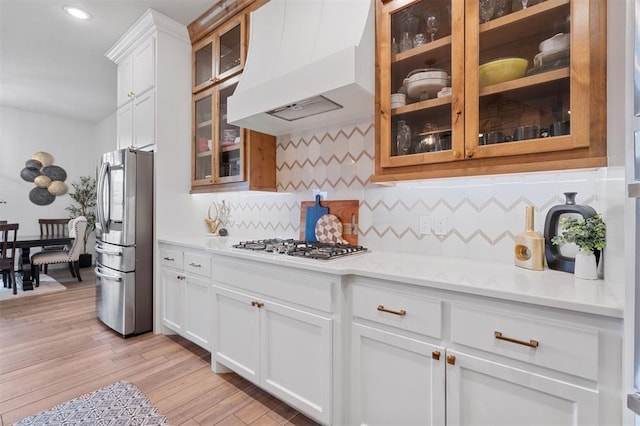 kitchen with light wood-type flooring, appliances with stainless steel finishes, custom range hood, decorative backsplash, and white cabinets