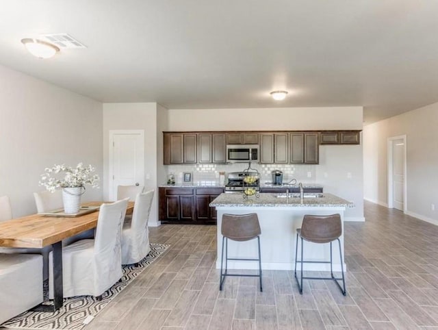 kitchen with light stone counters, dark brown cabinets, an island with sink, stainless steel appliances, and decorative backsplash