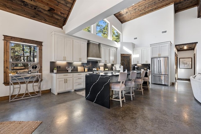 kitchen featuring wall chimney exhaust hood, a breakfast bar area, white cabinetry, wooden ceiling, and stainless steel fridge