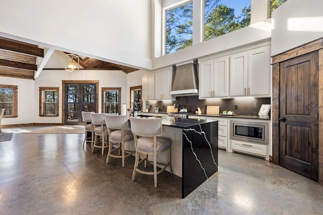 kitchen featuring a towering ceiling, stainless steel microwave, a kitchen bar, concrete floors, and wall chimney exhaust hood