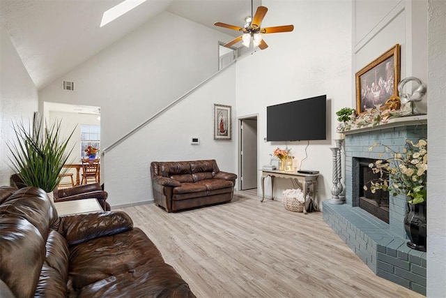 living room featuring high vaulted ceiling, a skylight, ceiling fan, light hardwood / wood-style floors, and a brick fireplace