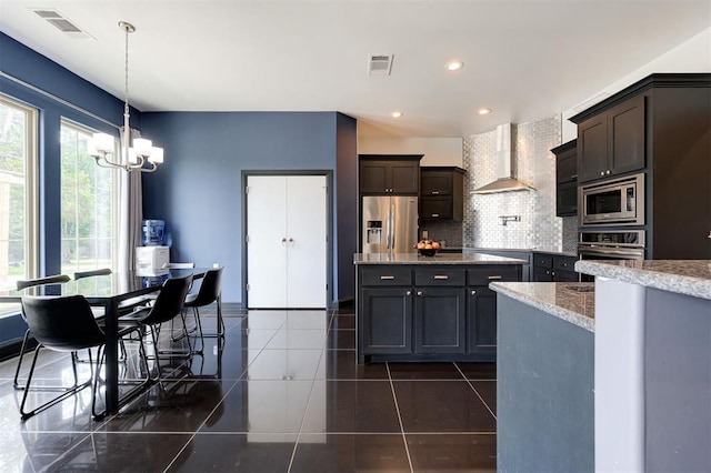 kitchen featuring wall chimney exhaust hood, a chandelier, appliances with stainless steel finishes, pendant lighting, and decorative backsplash