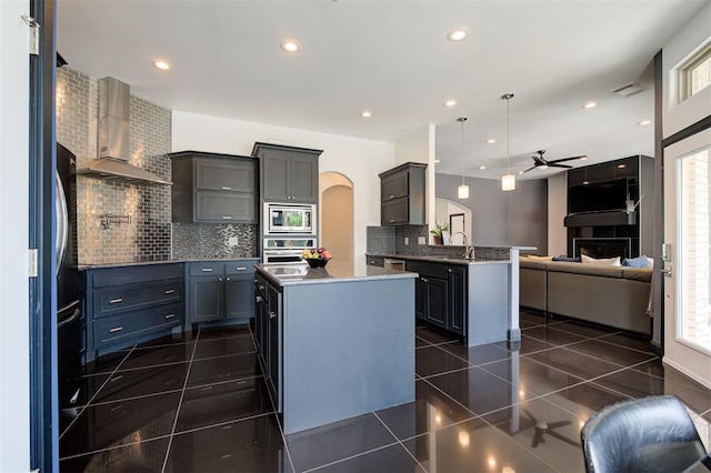 kitchen featuring wall chimney range hood, dark tile patterned floors, stainless steel appliances, a kitchen island, and decorative light fixtures