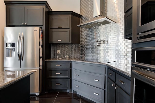 kitchen with wall chimney range hood, gray cabinets, appliances with stainless steel finishes, light stone counters, and decorative backsplash