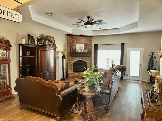 living room featuring visible vents, a ceiling fan, wood finished floors, a tray ceiling, and a brick fireplace