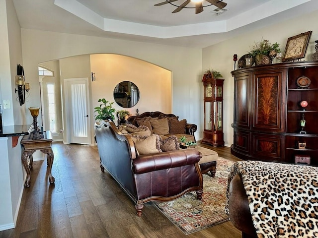 living area featuring ceiling fan, a raised ceiling, arched walkways, and dark wood finished floors