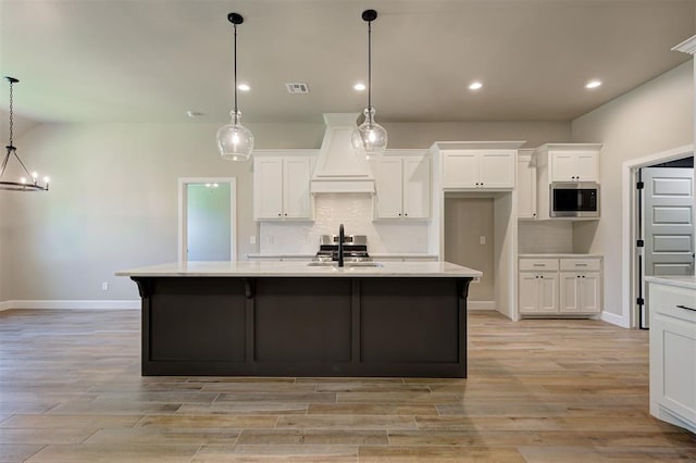 kitchen with custom exhaust hood, white cabinetry, decorative light fixtures, a center island with sink, and stainless steel appliances