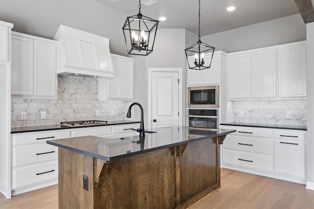 kitchen featuring a center island with sink, dark countertops, light wood-style flooring, appliances with stainless steel finishes, and custom exhaust hood