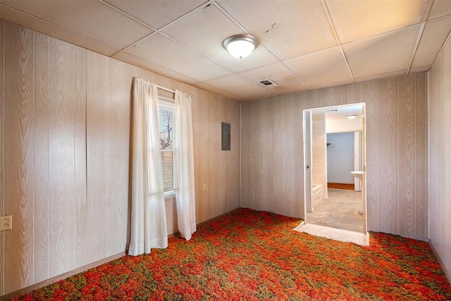 empty room with a paneled ceiling, carpet flooring, electric panel, and wood walls