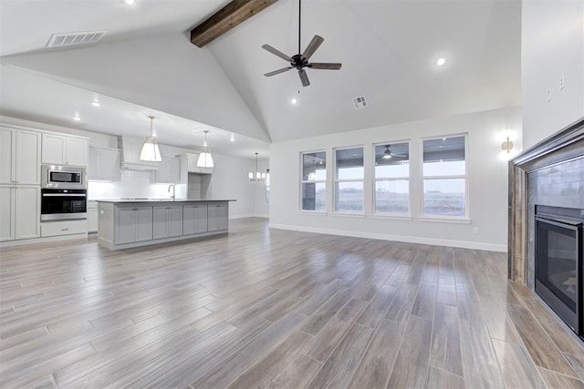 unfurnished living room featuring ceiling fan with notable chandelier, high vaulted ceiling, a tiled fireplace, light hardwood / wood-style floors, and beam ceiling