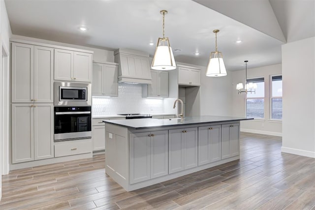 kitchen featuring backsplash, custom exhaust hood, hanging light fixtures, stainless steel appliances, and a center island with sink