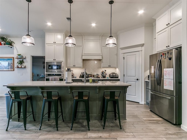 kitchen with custom exhaust hood, appliances with stainless steel finishes, a kitchen island with sink, and decorative light fixtures