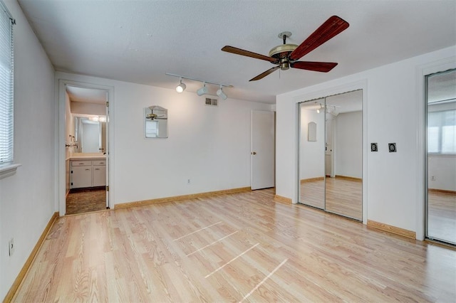 unfurnished bedroom with rail lighting, ensuite bath, light wood-type flooring, multiple closets, and ceiling fan