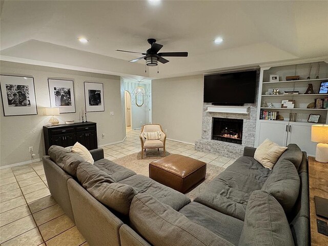 living room featuring ceiling fan, a tray ceiling, a stone fireplace, and light tile patterned floors