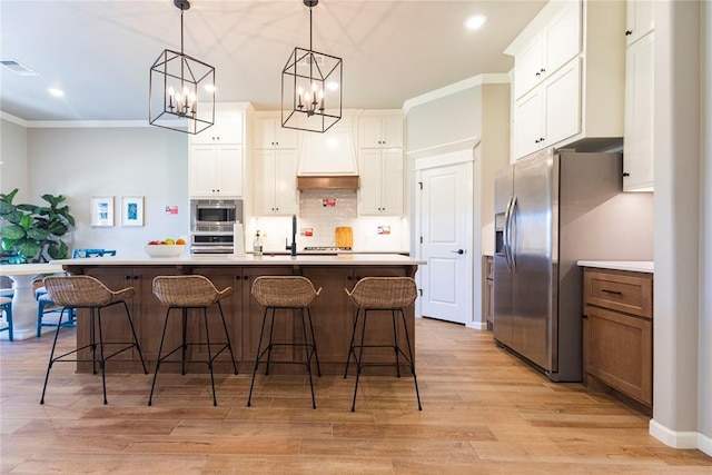 kitchen with premium range hood, white cabinetry, decorative light fixtures, appliances with stainless steel finishes, and an island with sink