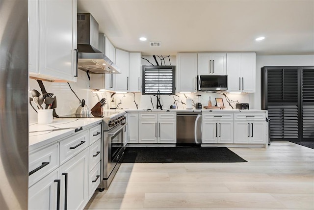 kitchen featuring appliances with stainless steel finishes, white cabinetry, light stone counters, wall chimney range hood, and light hardwood / wood-style flooring