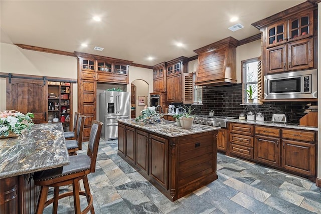 kitchen with light stone counters, a center island with sink, custom range hood, stainless steel appliances, and a barn door