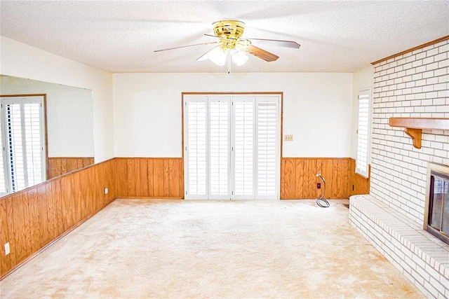 unfurnished living room with ceiling fan, a textured ceiling, light carpet, a brick fireplace, and wood walls