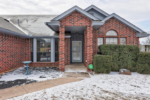 snow covered property entrance with roof with shingles and brick siding