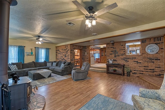 living room featuring wood-type flooring, a textured ceiling, a wood stove, ceiling fan, and brick wall