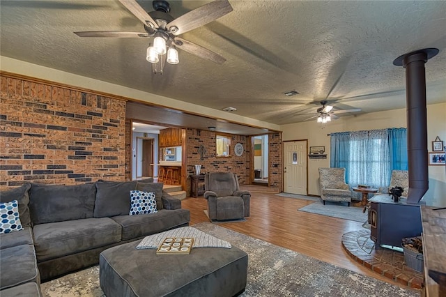 living room featuring a textured ceiling, a wood stove, hardwood / wood-style flooring, ceiling fan, and brick wall