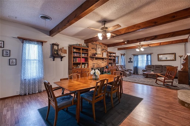 dining area featuring ceiling fan, beam ceiling, a fireplace, wood-type flooring, and a textured ceiling