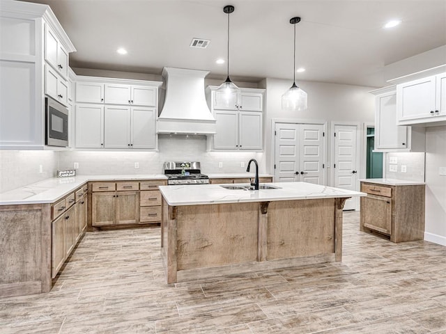 kitchen with white cabinets, custom range hood, appliances with stainless steel finishes, and a center island with sink