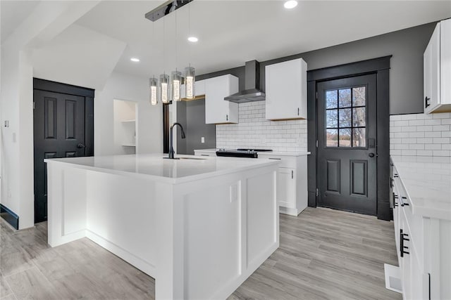 kitchen featuring white cabinets, a kitchen island with sink, hanging light fixtures, and wall chimney range hood