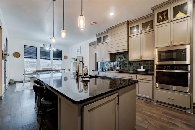 kitchen with sink, appliances with stainless steel finishes, a kitchen island with sink, hanging light fixtures, and dark stone countertops