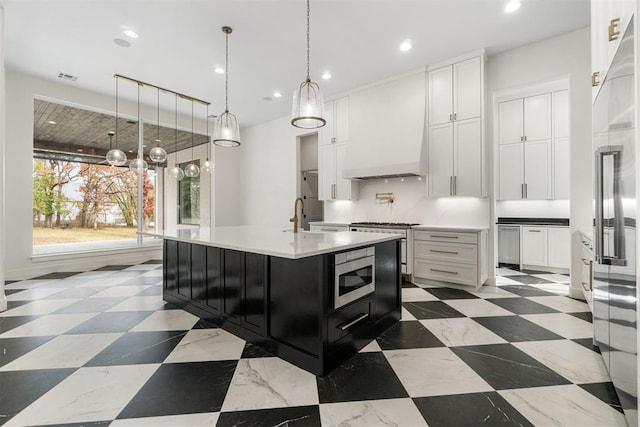 kitchen featuring premium range hood, appliances with stainless steel finishes, an island with sink, pendant lighting, and white cabinets