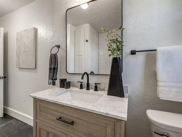 bathroom featuring toilet, a textured wall, vanity, and baseboards