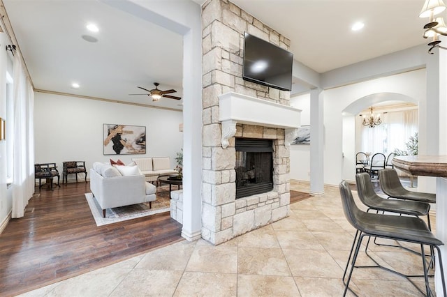 living room featuring light tile patterned flooring, ornamental molding, a stone fireplace, and ceiling fan with notable chandelier